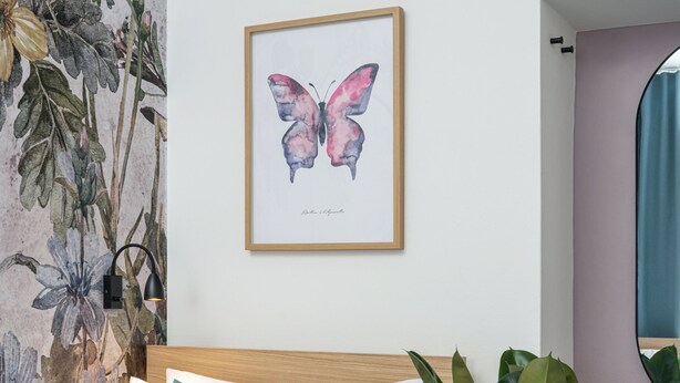 Una cama con cabecera de madera, un almohadón azul y una almohada blanca. En la pared, un cuadro enmarcado con una mariposa.