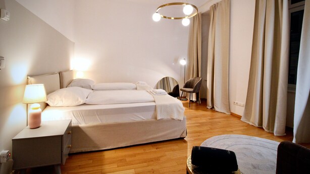 A hotel room with a double bed, a bedside table with a lamp, a chair, and a large window with curtains. - Stylish Home in Vienna’s Central 9th District (Vienna)