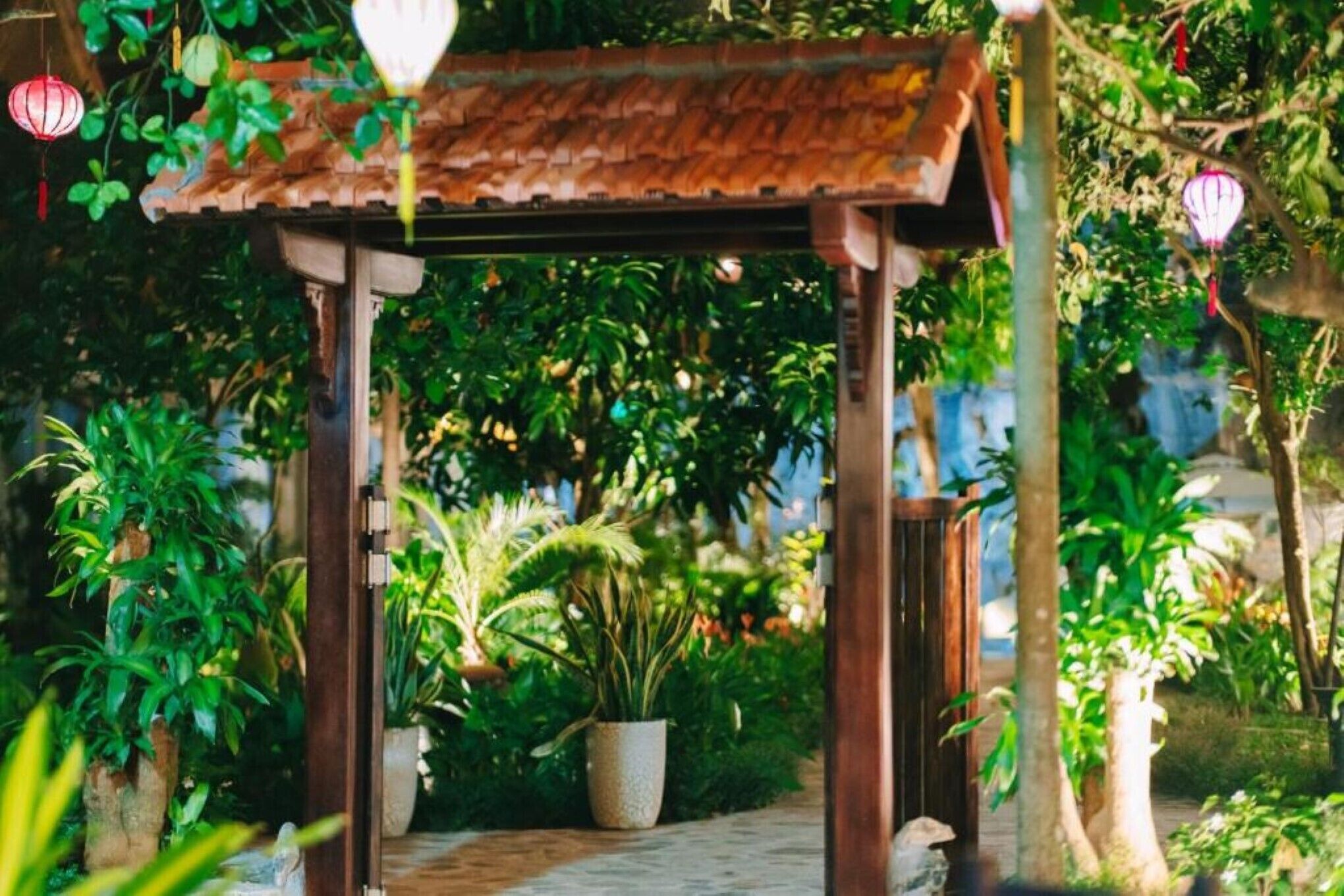 Un gazebo in legno con tetto in tegole, immerso nel verde e circondato da piante in vaso.