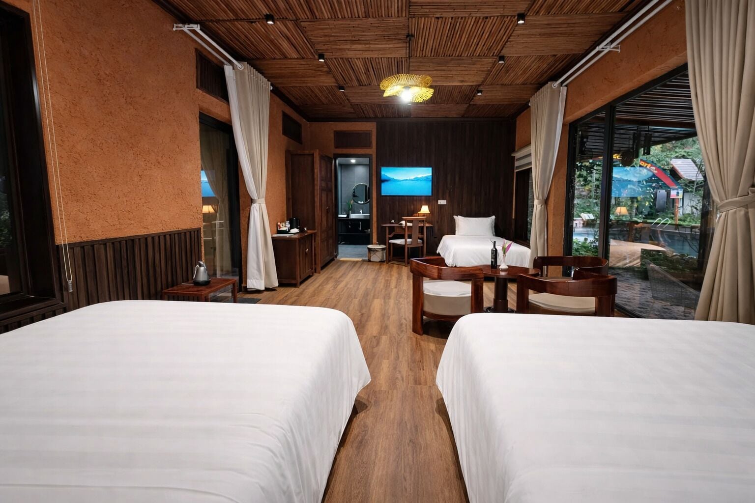 A hotel room with two beds, wooden flooring, a TV, a desk, and a view of the outdoors.