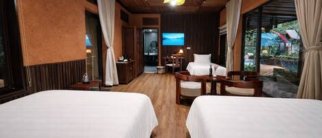 A hotel room with two beds, wooden flooring, a TV, a desk, and a view of the outdoors.
