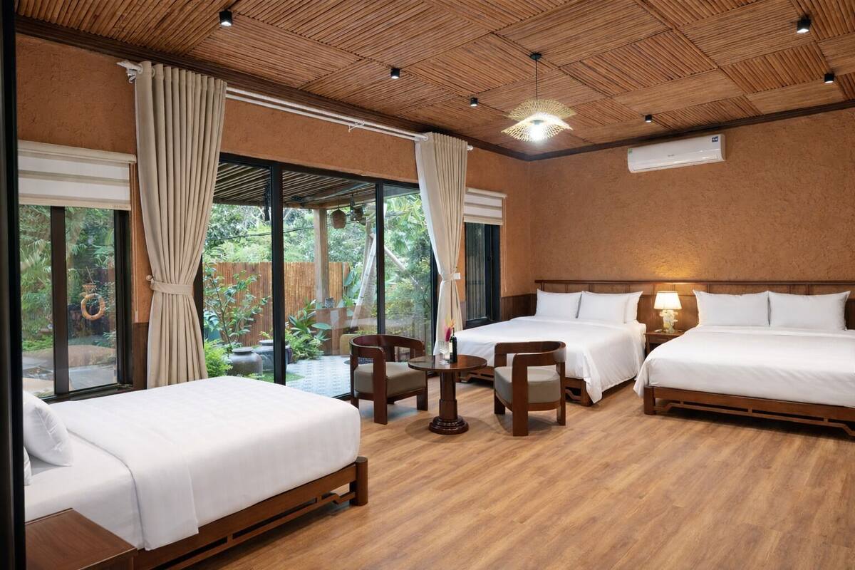 Quarto de hotel com duas camas, uma mesa pequena e cadeiras. Há uma janela grande com cortinas e um aparelho de ar condicionado na parede.