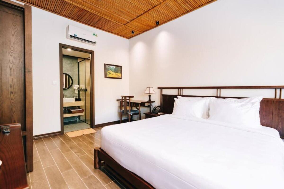 Quarto de hotel com teto de madeira, cama com roupa de cama branca, pequena mesa de trabalho com abajur e banheiro com porta de vidro.
