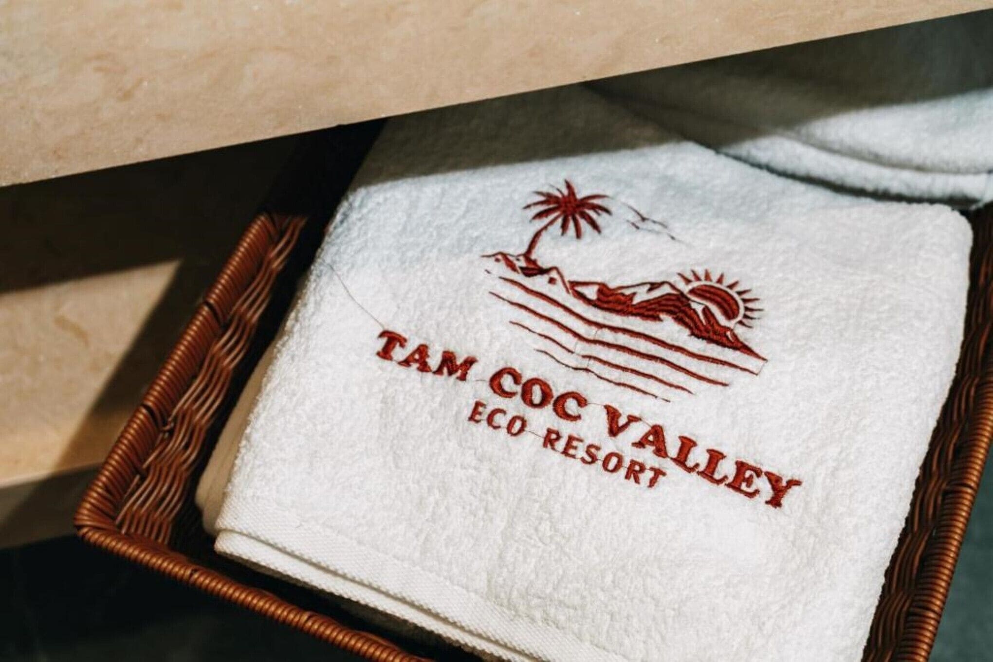 Una toalla blanca con el bordado "TAM COC VALLEY ECO RESORT", sobre una bandeja de mimbre.