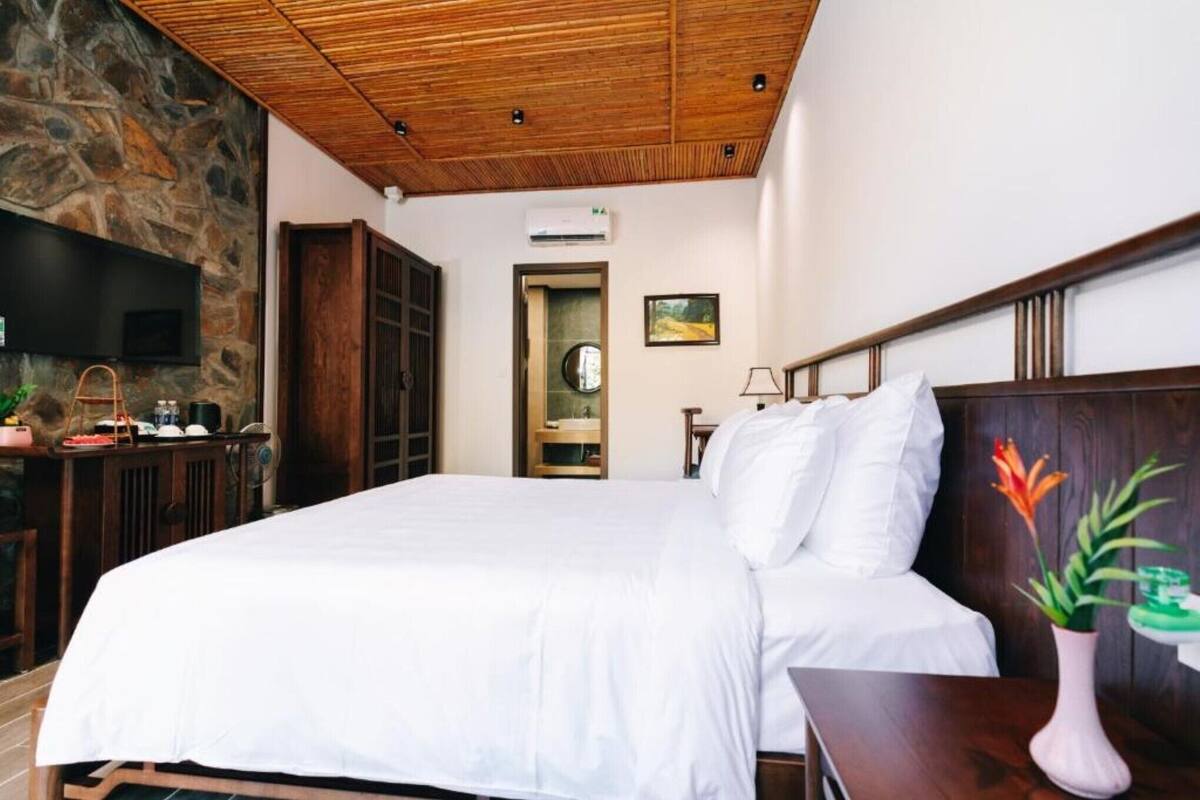 Quarto com cama grande, cabeceira de madeira, parede de pedra e teto com vigas de madeira.