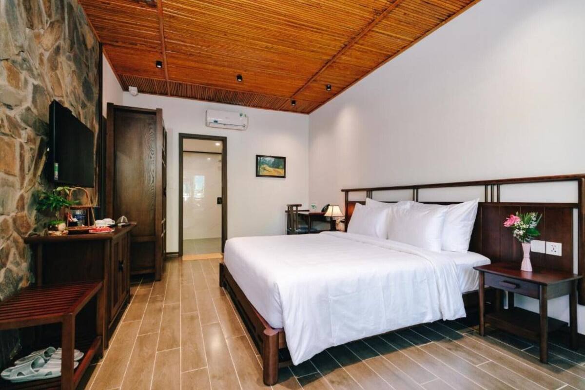 Quarto com teto de madeira, parede de pedra, cama com roupa branca, mesa de cabeceira, escrivaninha e uma cadeira.