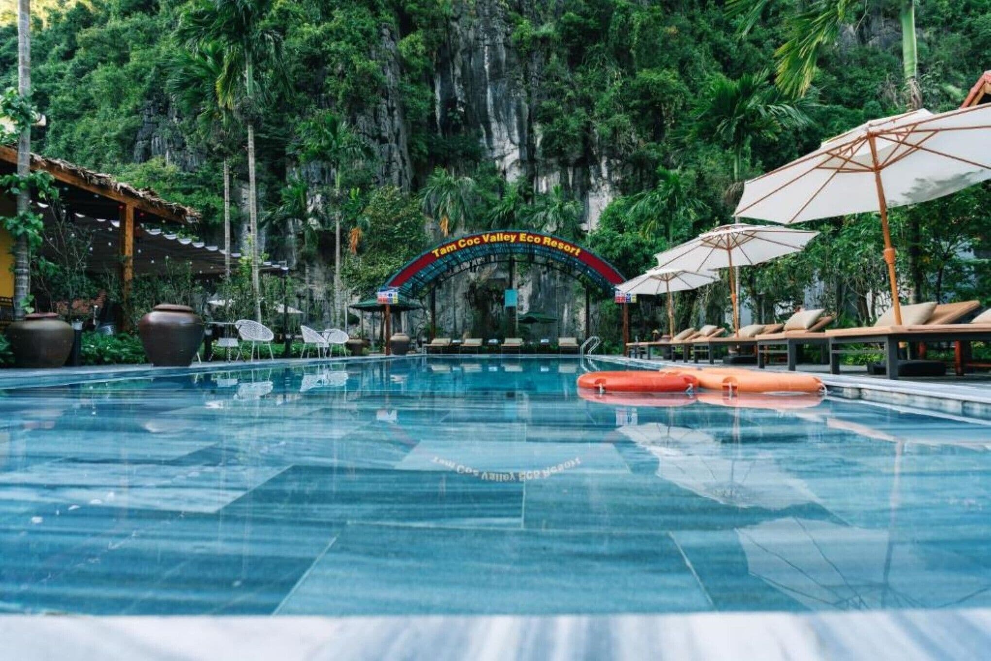 Una piscina con sillas de descanso y sombrillas, rodeada de abundante vegetación y un telón de fondo rocoso.