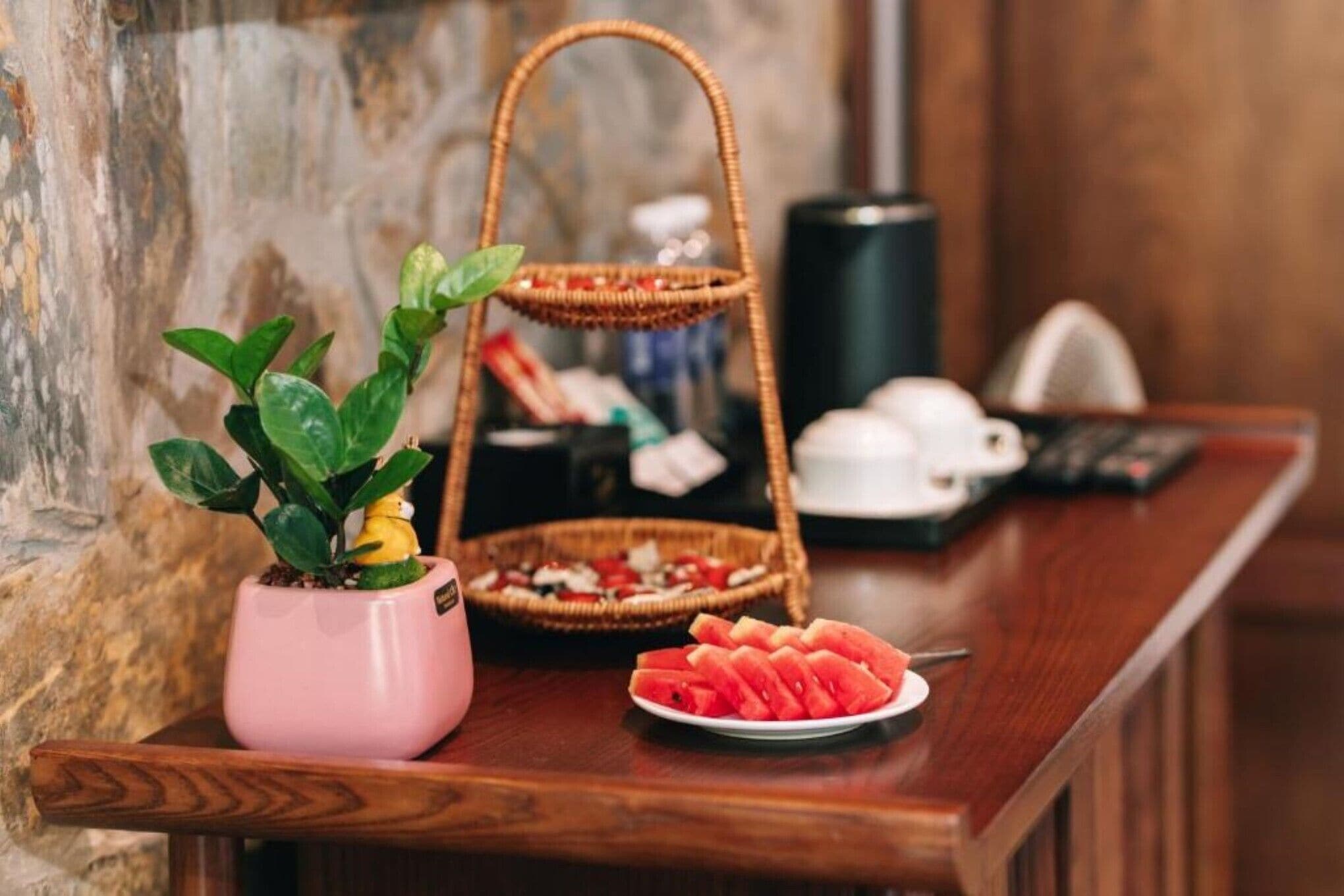 A wooden table with a pink potted plant, a basket with snacks, and a plate of watermelon slices.