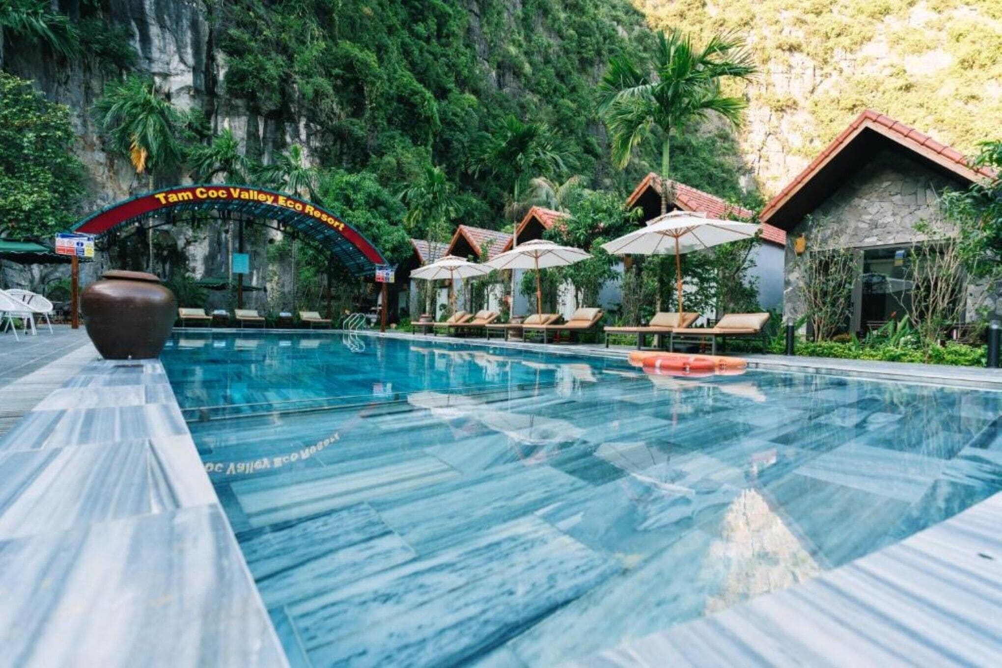 A pool area with lounge chairs and umbrellas, surrounded by lush greenery and a mountainous backdrop.