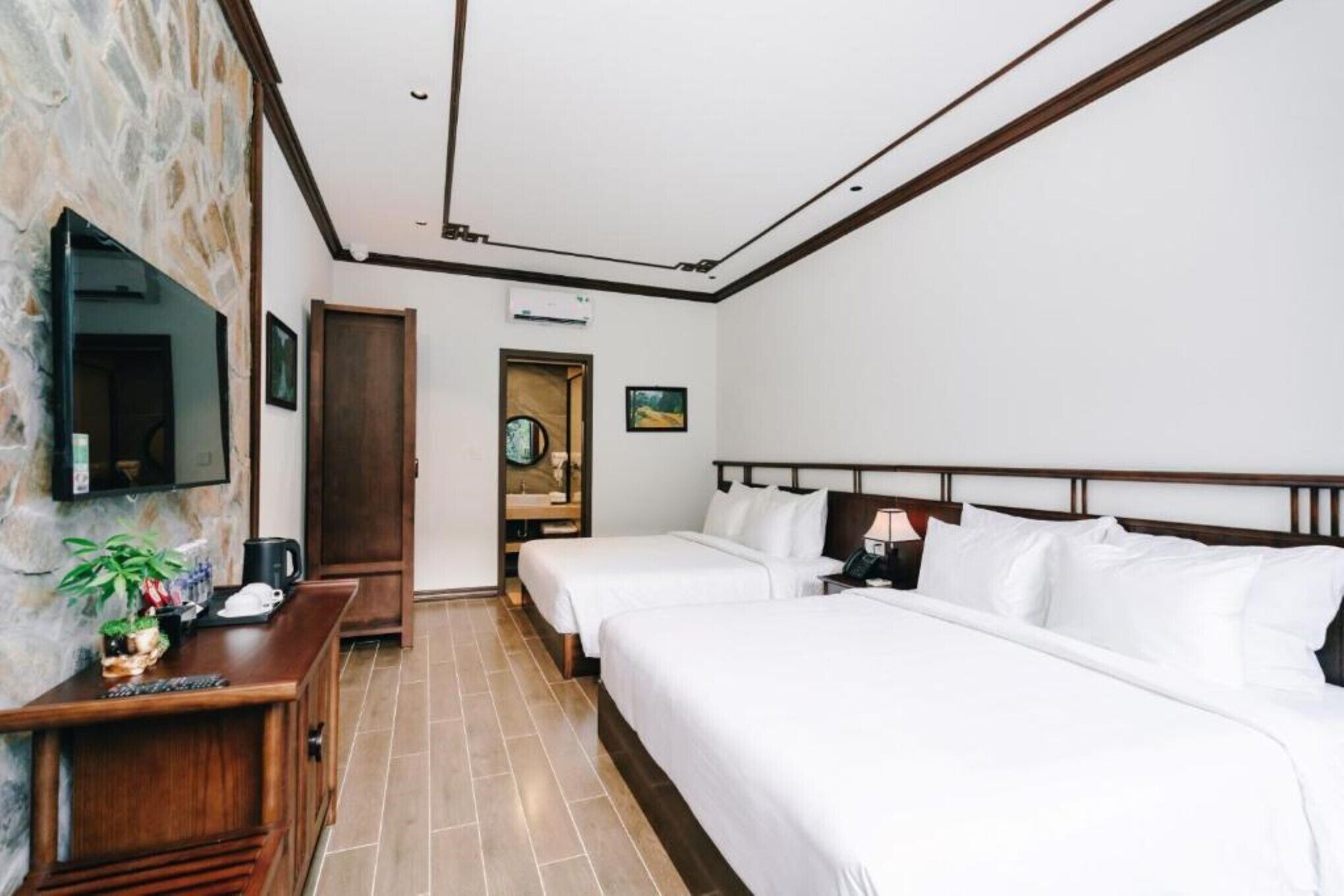 Une chambre d’hôtel avec deux lits simples, un bureau en bois agrémenté d’une plante, un téléviseur à écran plat et un mur en pierre.