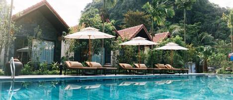 Un área de piscina con sillones de descanso y sombrillas, rodeada de vegetación exuberante y montañas al fondo.