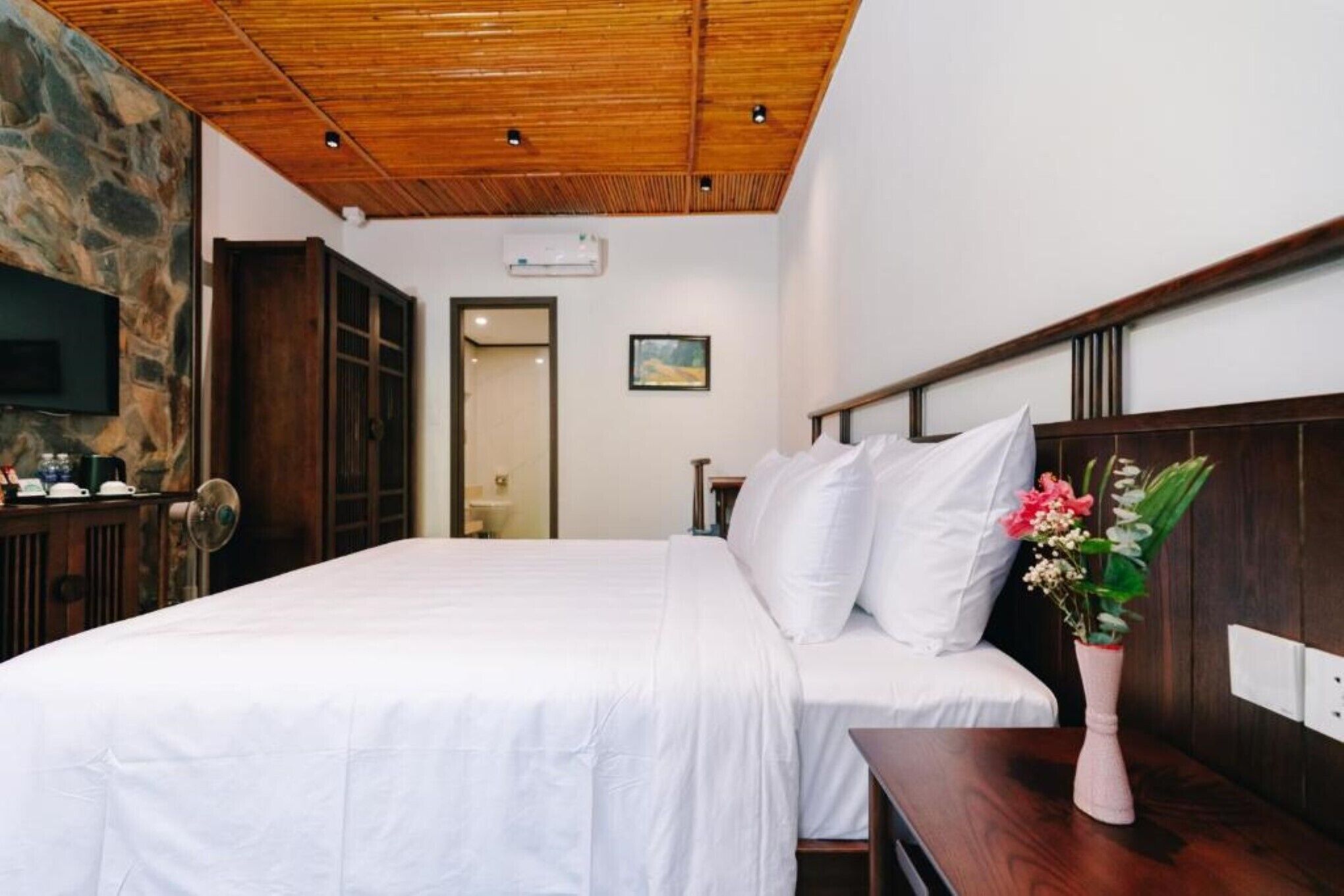 A bedroom with a wooden ceiling, a stone wall, a bed with white bedding, a wooden nightstand, a vase with flowers, and a television mounted on the wall.