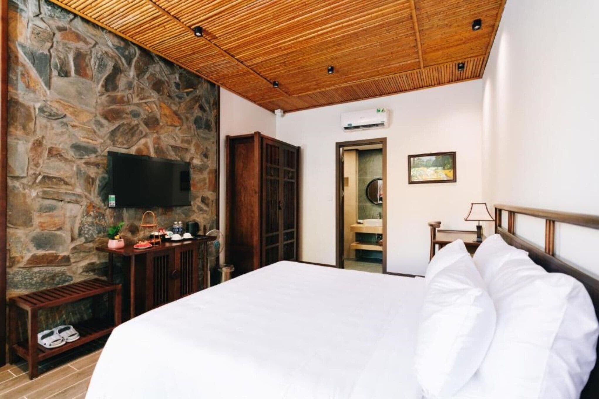 A bedroom with a stone wall, a wooden ceiling, a bed with white bedding, a TV mounted on the wall, and a wooden cabinet with toiletries.