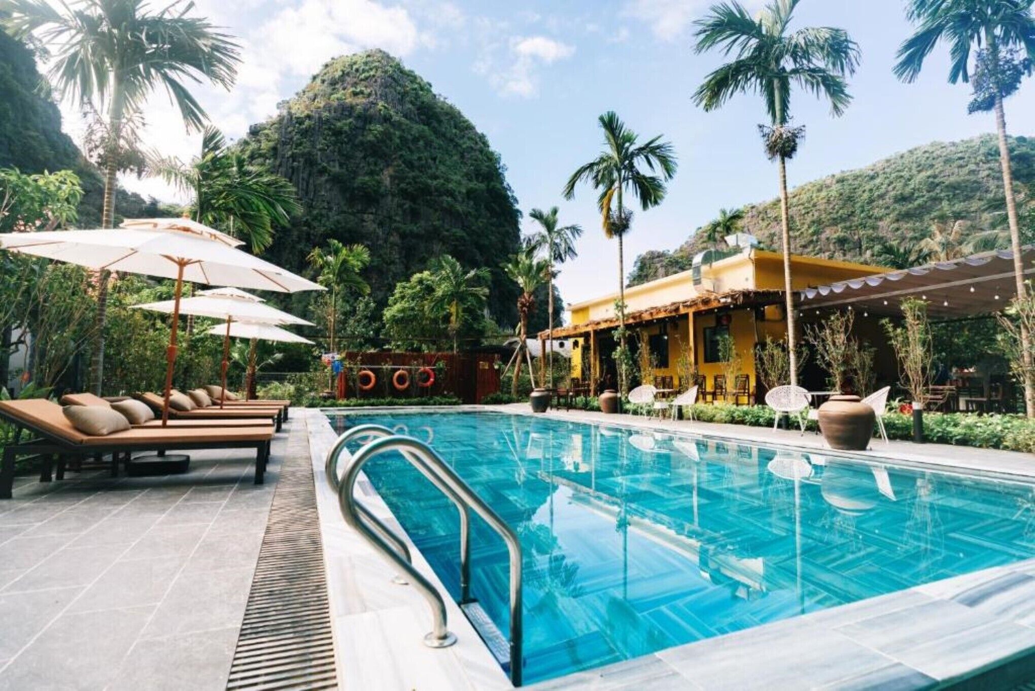 Zona de piscina con sillones de descanso y sombrillas, rodeada de palmeras y con una montaña al fondo.