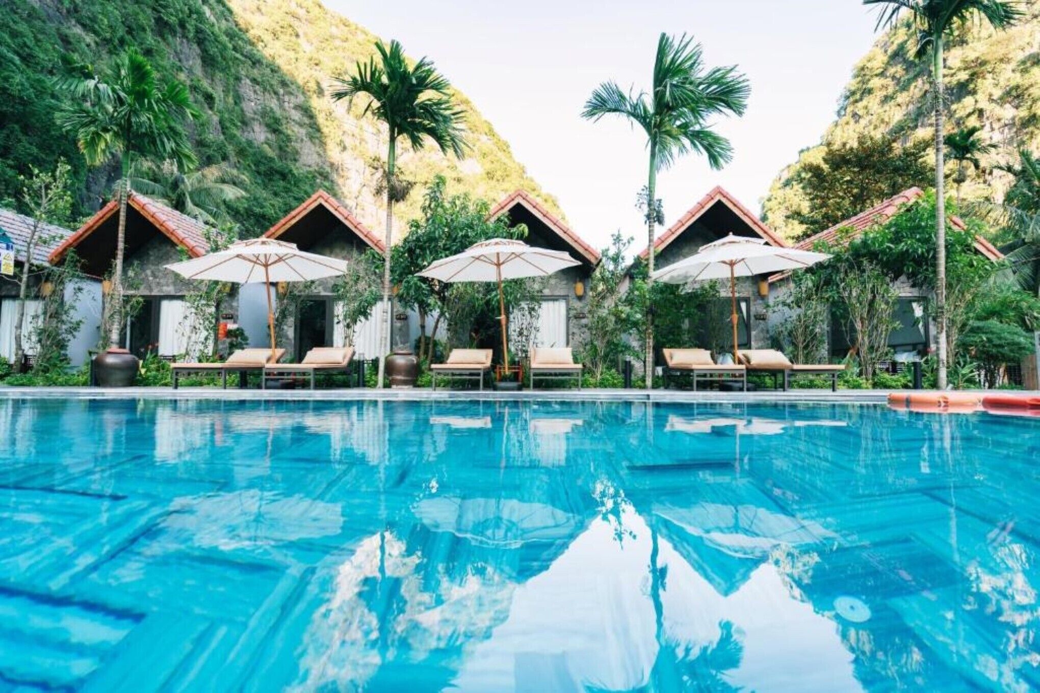 A pool with lounge chairs and umbrellas, surrounded by buildings with red roofs and greenery.