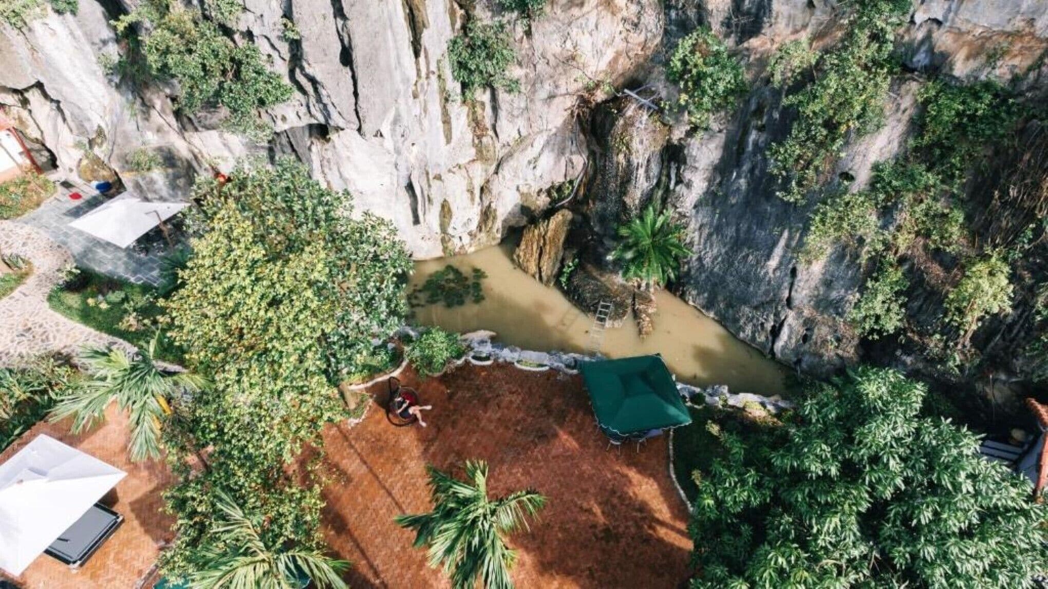 An aerial view of a cliffside area with a small pool, palm trees, and a green-roofed structure.