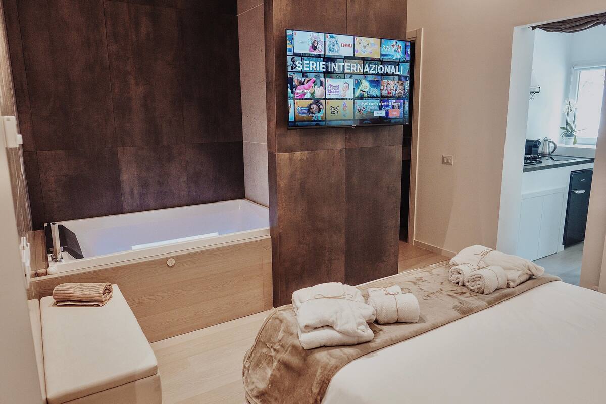 A hotel room with a large bed, a bathtub, a TV displaying international series, and a view of the kitchen.