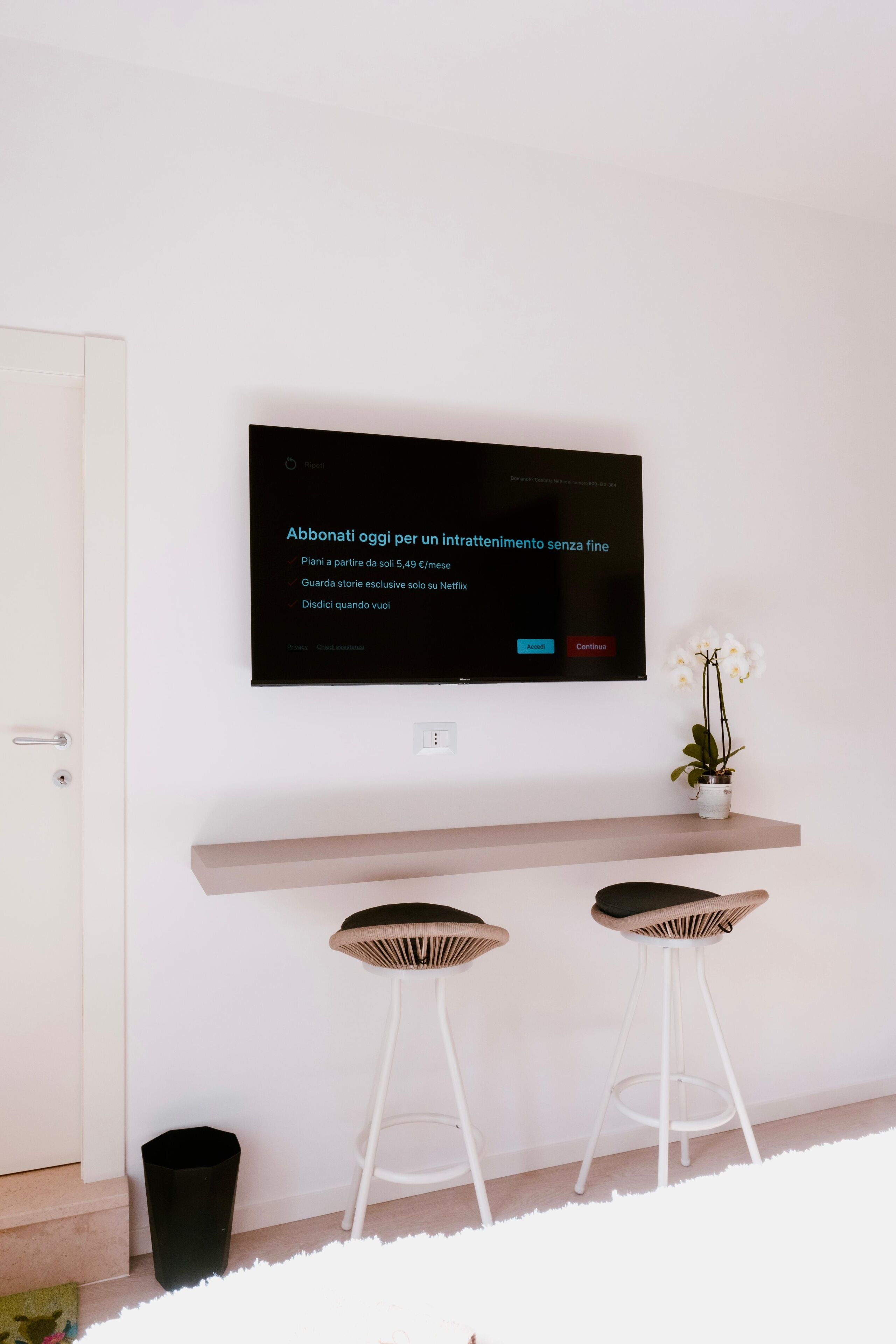Una sala moderna con televisor montado, dos taburetes, un estante pequeño con una planta y una puerta blanca.