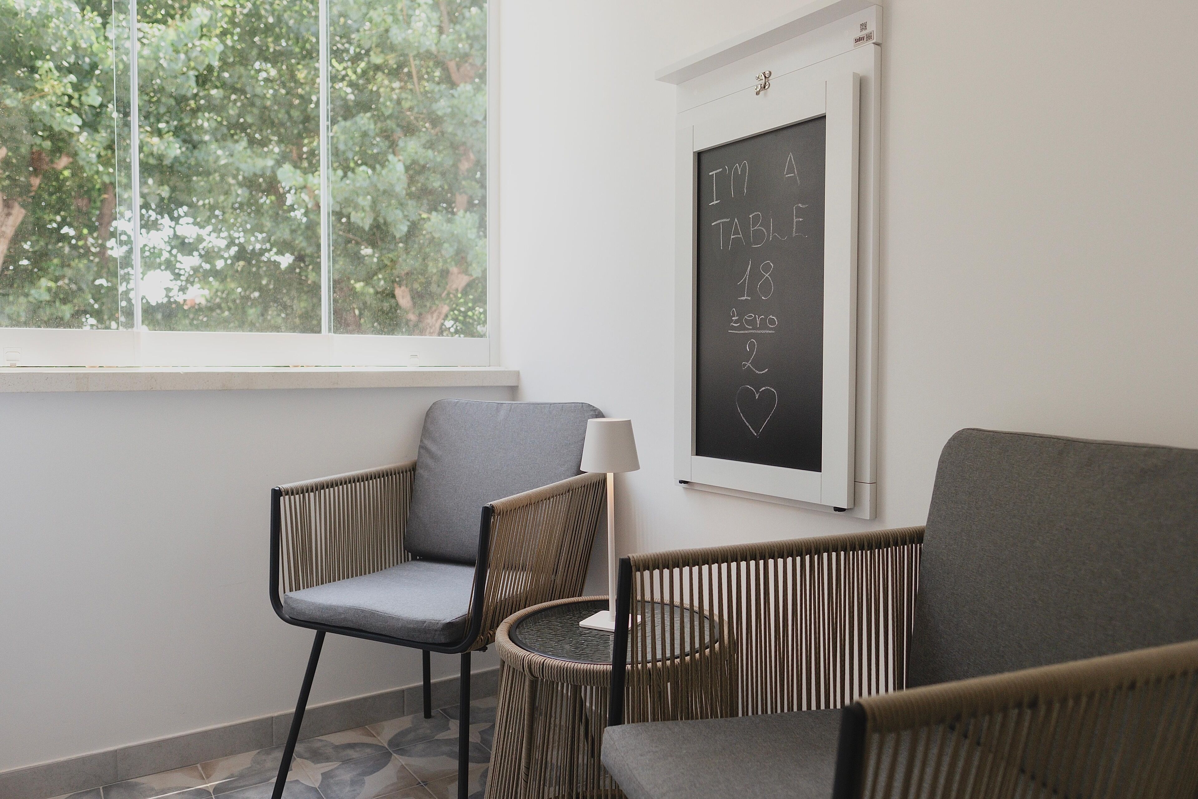 A room with a window, two chairs, a small table, and a wall-mounted blackboard with writing.