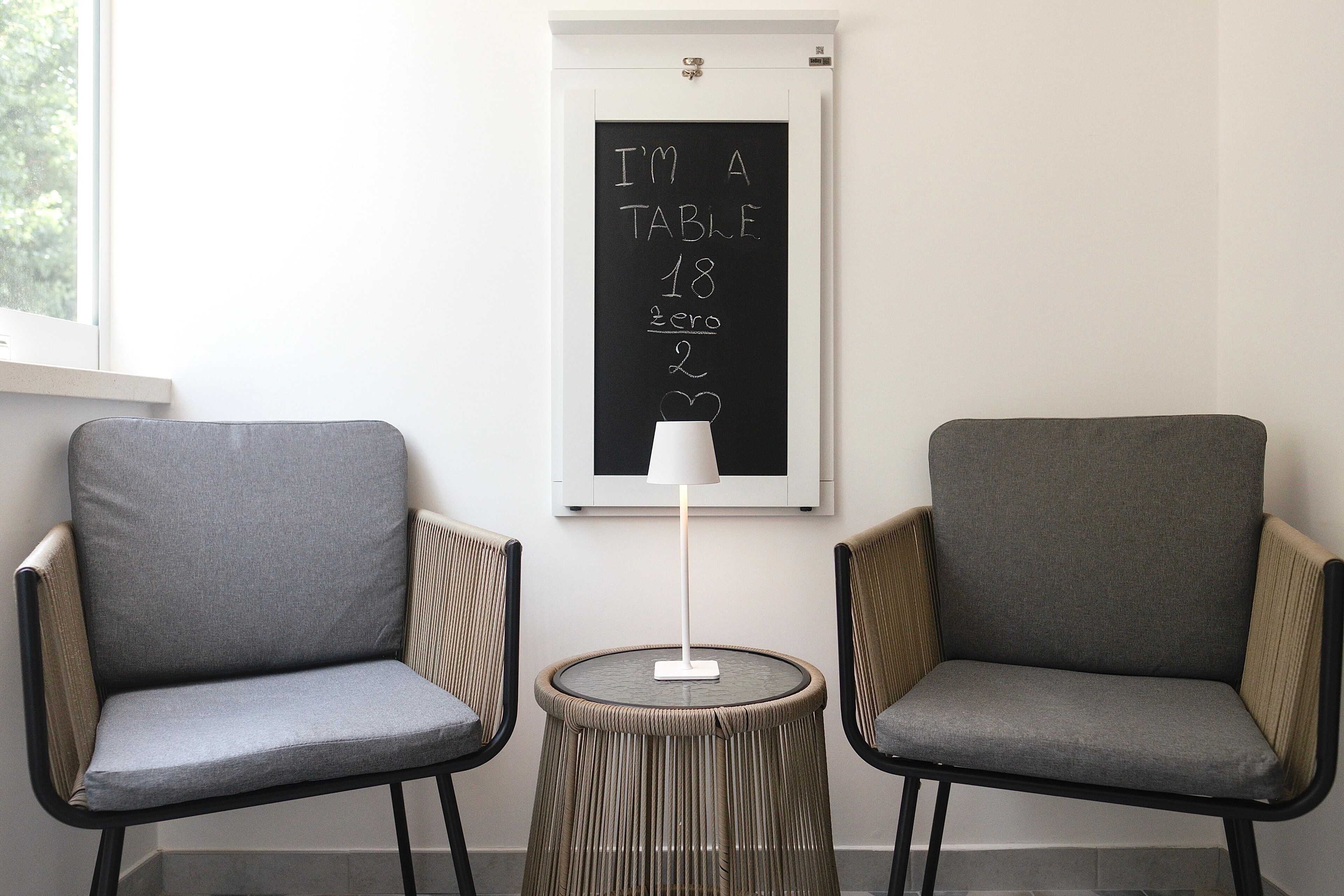 Two grey armchairs, a small round table with a lamp, and a wall-mounted blackboard with a chalk drawing.