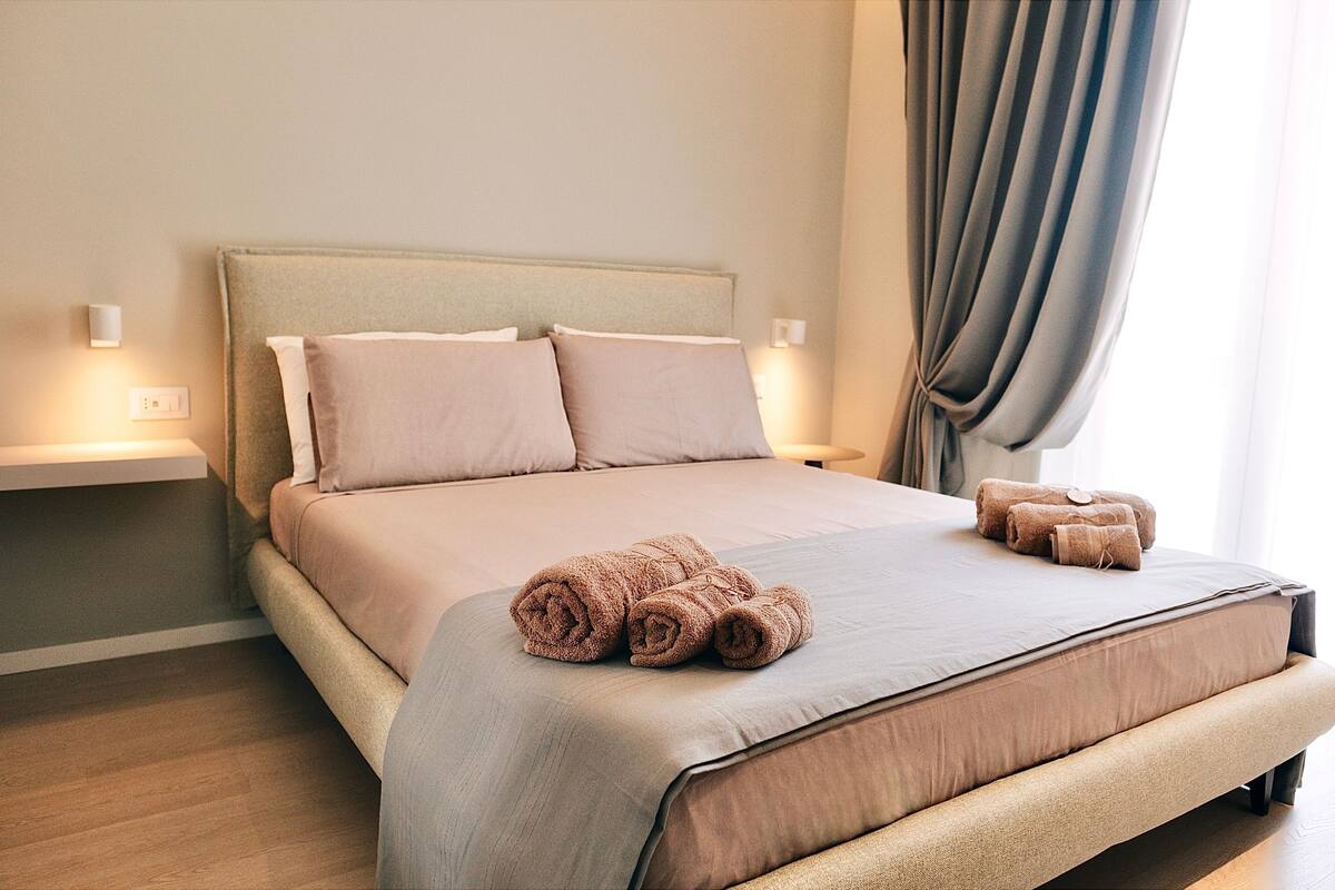 A neatly made bed with a beige bedspread and pillows, a bedside table with a lamp, and a window with curtains.