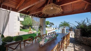 An outdoor dining area with a long wooden table and chairs, surrounded by stone walls and greenery.