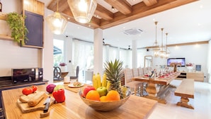 A modern kitchen with a wooden island, a dining area with a long table, and a television in the background.
