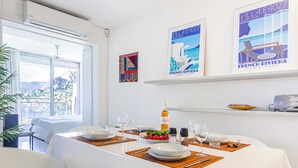 Zona pranzo con tavolo apparecchiato per due, con bottiglia di vino, bicchieri e una ciotola di frutta. Due sedie, una pianta in vaso e una finestra vista paesaggio.