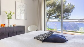 Camera da letto con un grande letto, vista sul mare, una sedia bianca e un tavolino con un vaso e frutta.