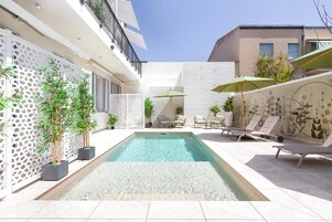 Area piscina moderna all'aperto con lettini e ombrelloni, circondata da piante in vaso e un muro decorativo.