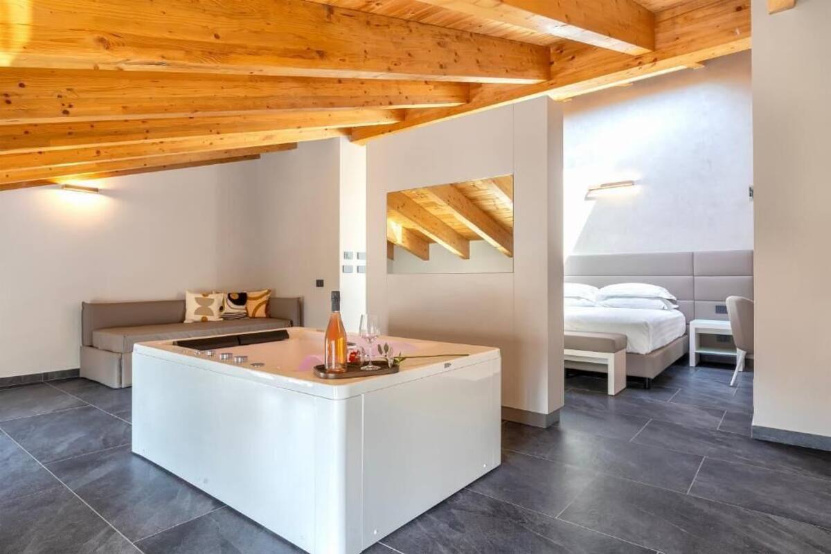 A modern hotel room with a wooden ceiling, a central island with a bottle and glasses, a sofa, a bed, and a mirror on the wall.
