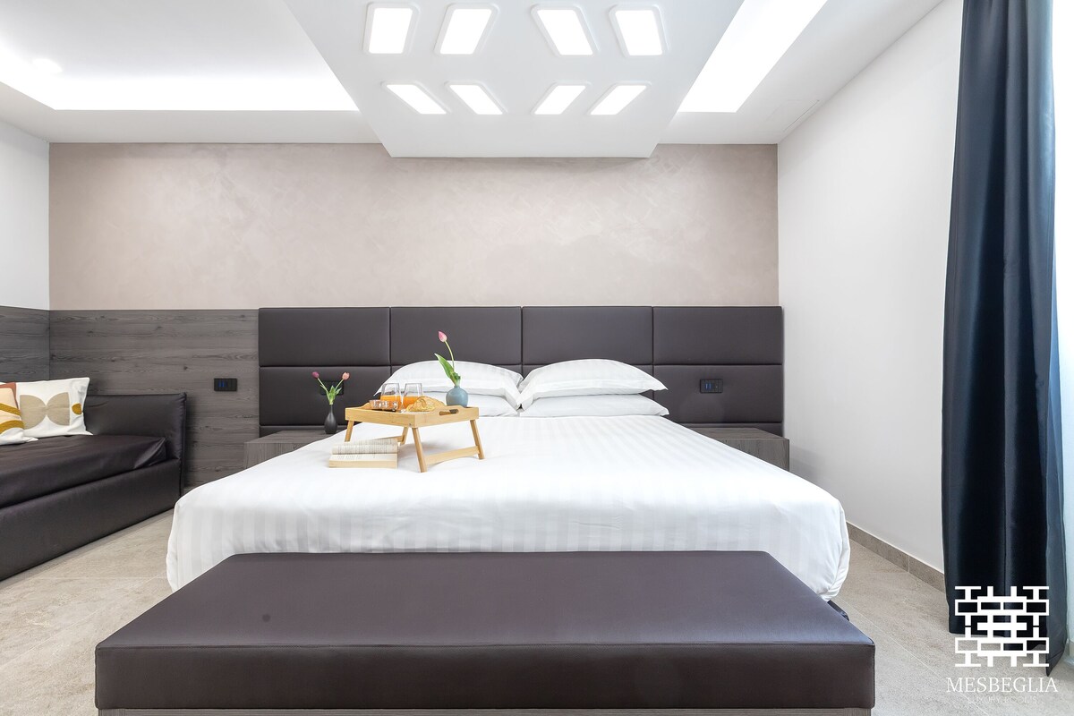 A modern hotel room with a large bed, a bedside table with a tray of breakfast, a dark sofa, and a large window with a curtain.