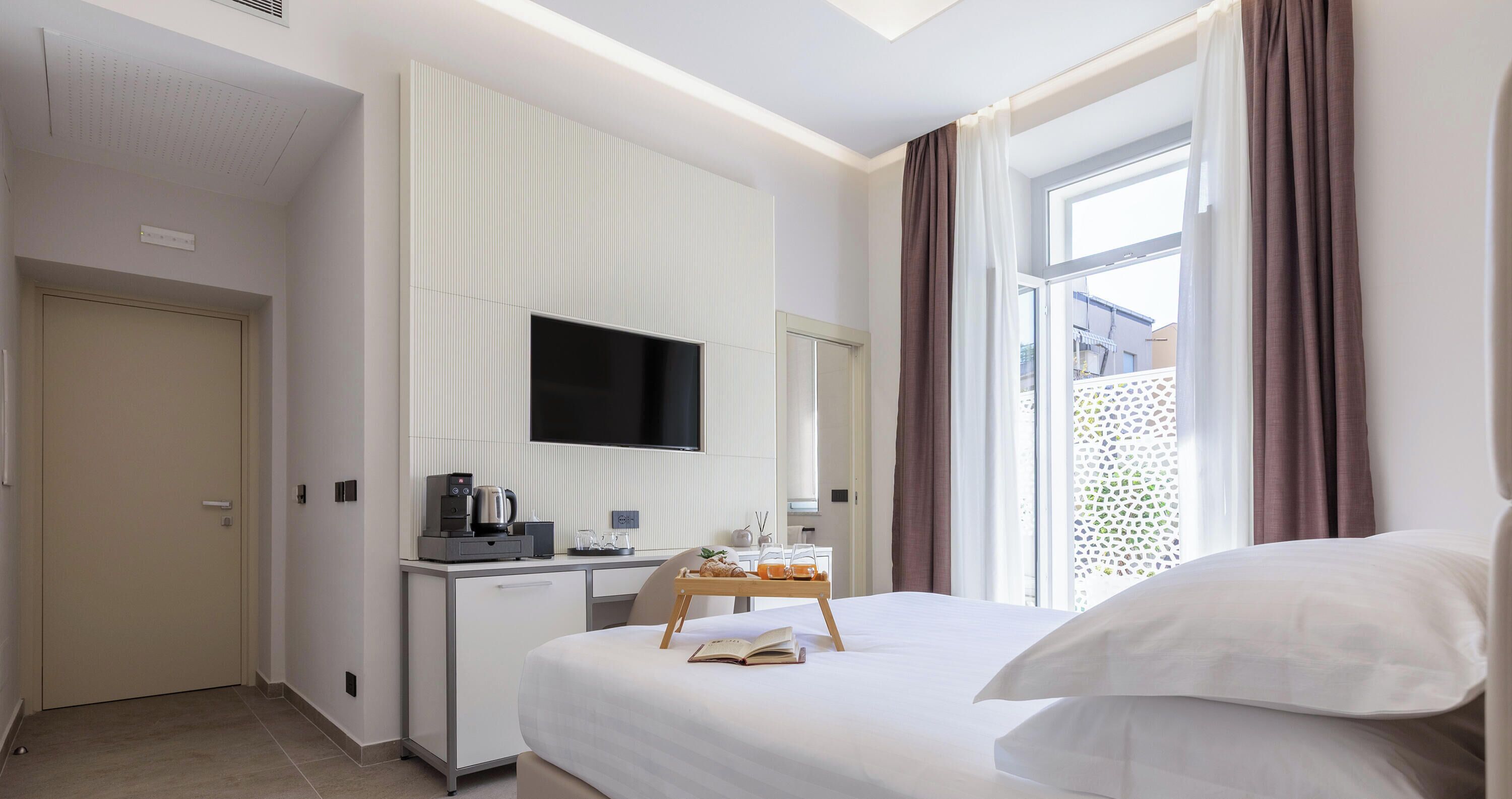 A hotel room with a bed, a desk with a flat-screen TV, a coffee machine, and a window with curtains.