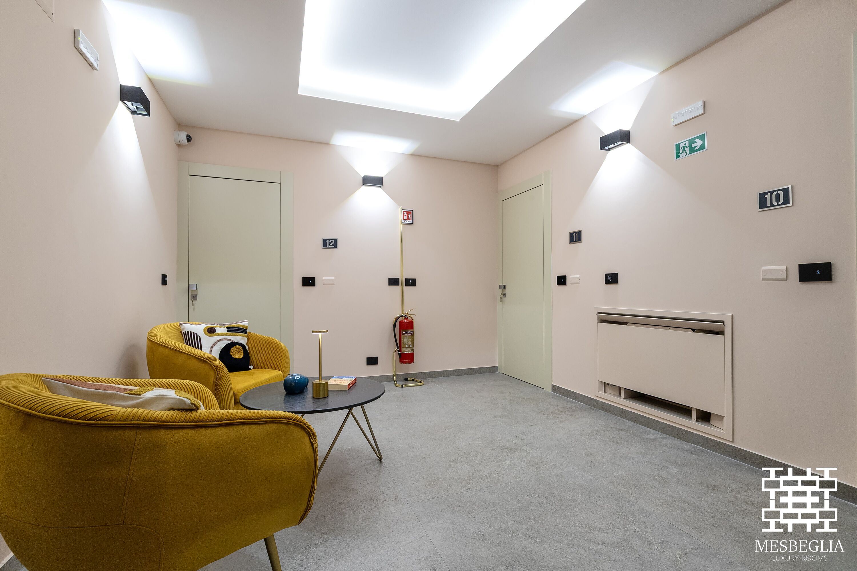 A hotel room with a yellow armchair, a small round table, a mirror, and a fire extinguisher.