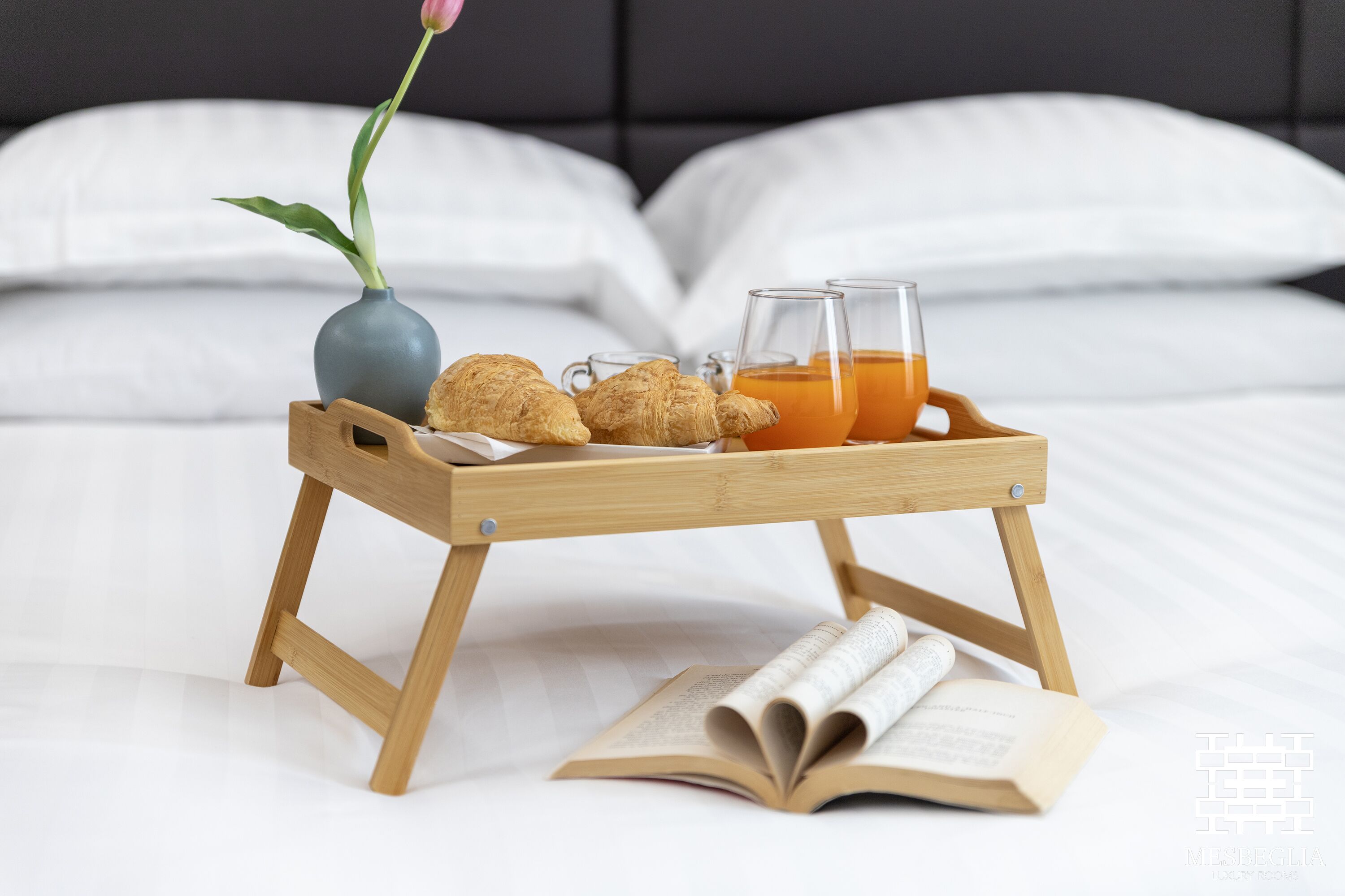 Una bandeja de madera con croissants, un florero con una flor y vasos de jugo de naranja sobre una cama.