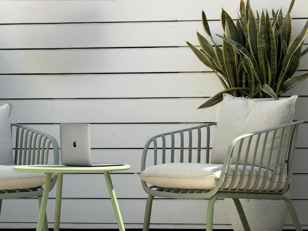 Eine Terrassenfläche mit zwei Stühlen, einem kleinen Tisch, einer Topfpflanze und einem Laptop an einer weißen Wand.
