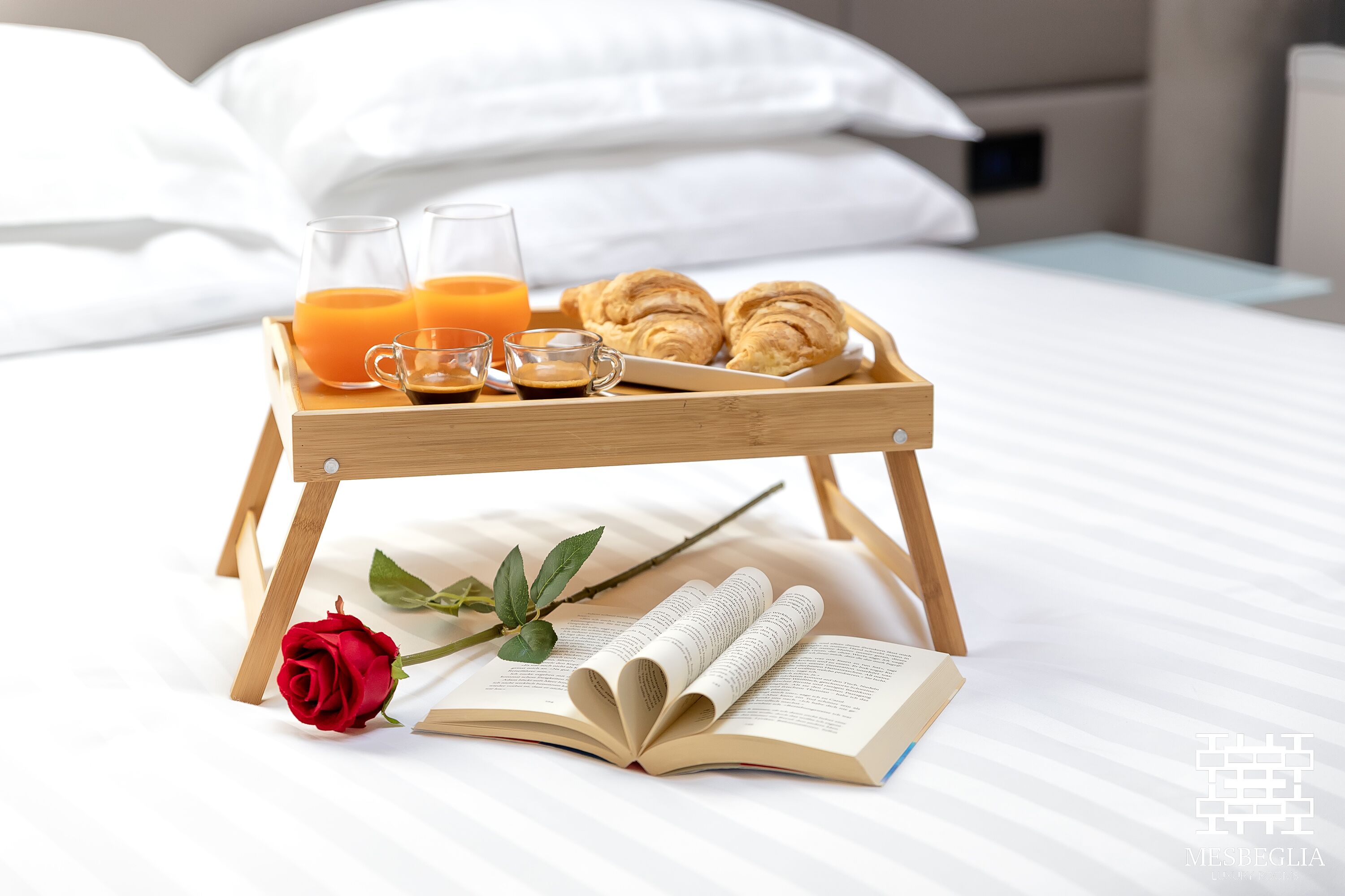 A wooden tray with croissants, juice, and coffee on a bed with white bedding.