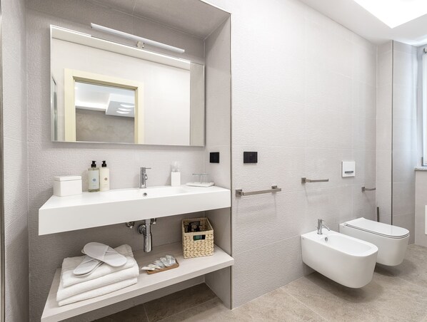 Un baño moderno con un espejo grande, dos lavamanos, un bidé y un inodoro. Hay un perchero y un estante con artículos de aseo.