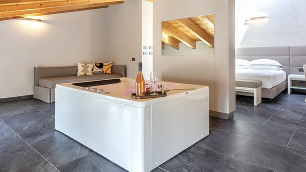 A modern, spacious room with a central island, a sofa, a bed, and a small table. - MESBEGLIA LUXURY ROOMS (Alghero)