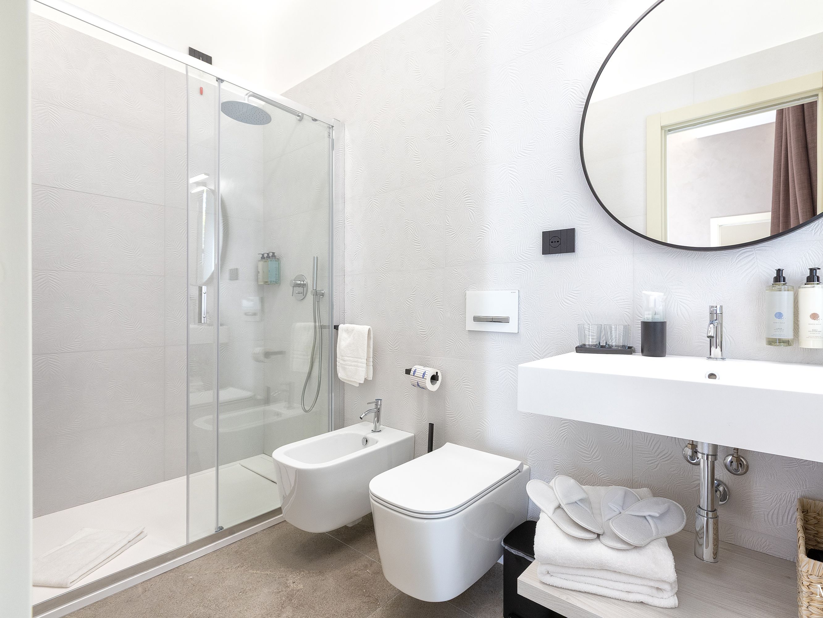 Une salle de bain moderne avec une douche à l’italienne en verre, un bidet, des toilettes, un lavabo avec un miroir et des serviettes soigneusement pliées.