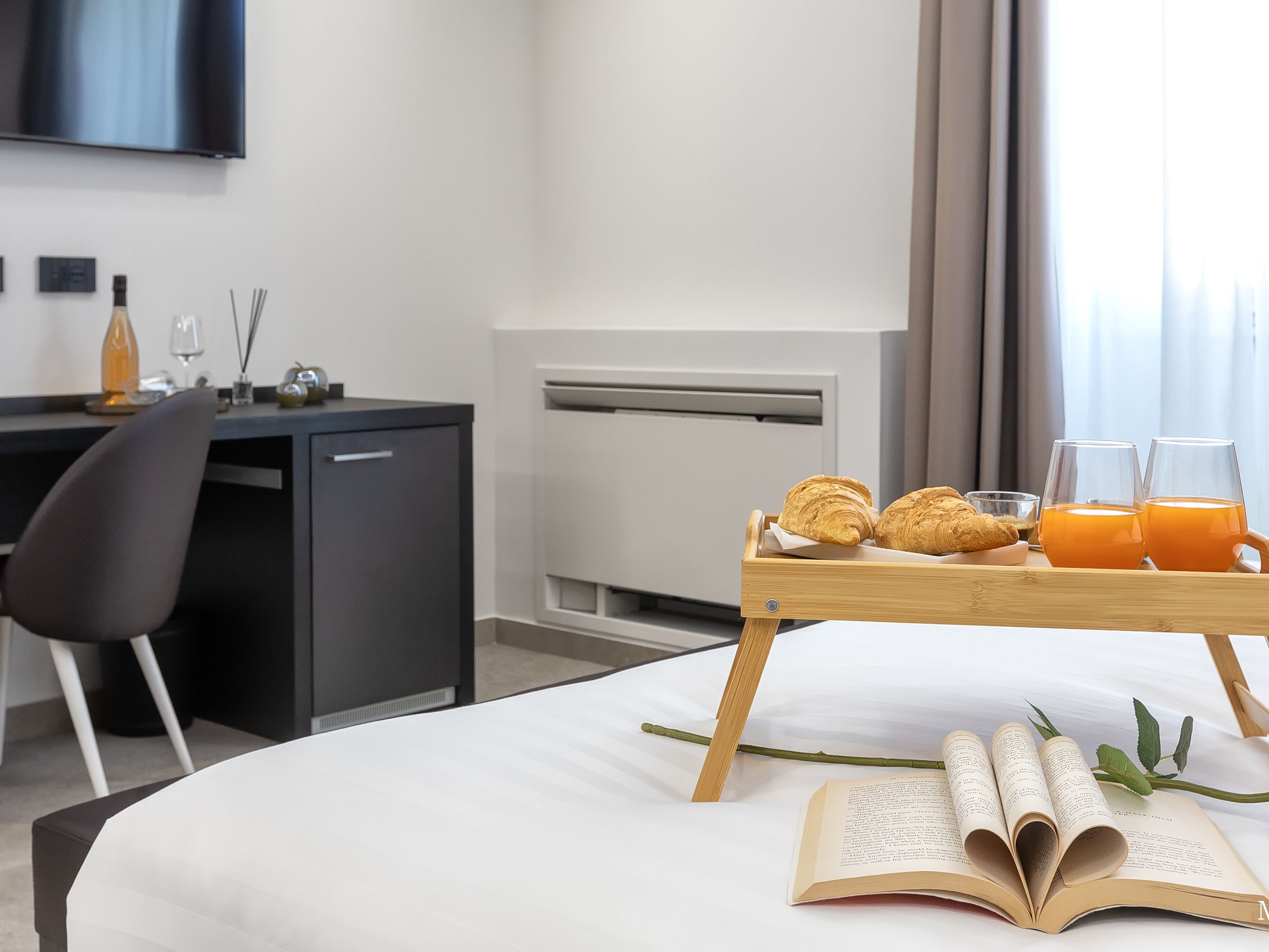 Una cama bien tendida con una bandeja de cruasanes, zumo de naranja y un libro, un escritorio con una botella y vasos, y un televisor montado en la pared.