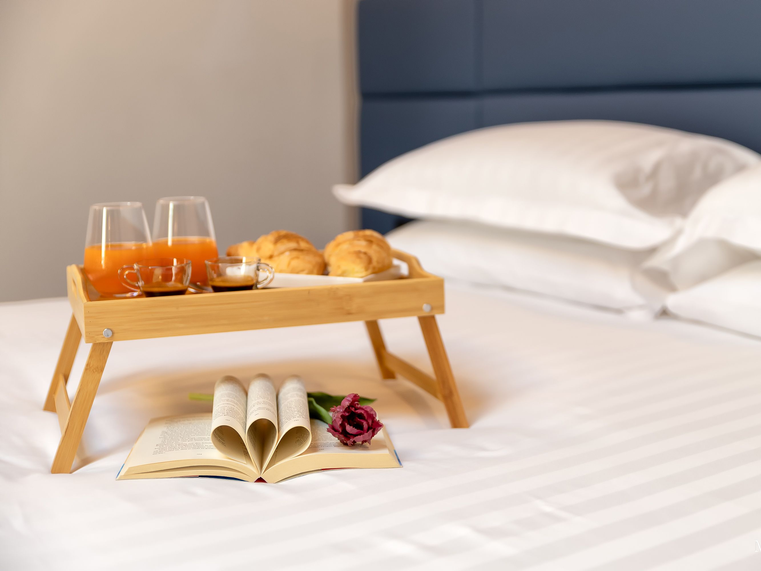A neatly made bed with a wooden tray holding breakfast items, a book, and a rose.