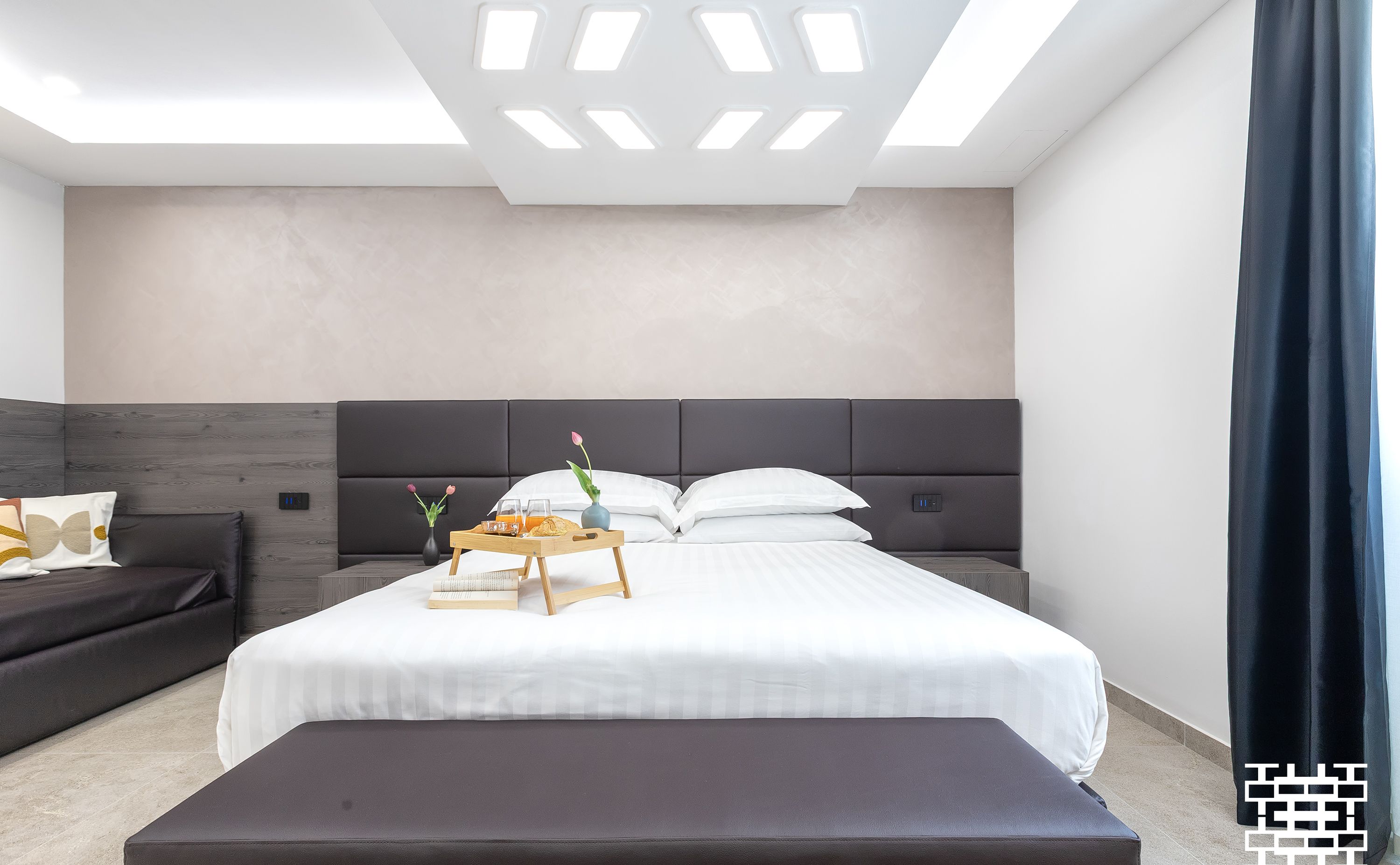 A modern hotel room with a large bed, a bedside table with a tray of breakfast, a dark sofa, and a large window with a curtain.