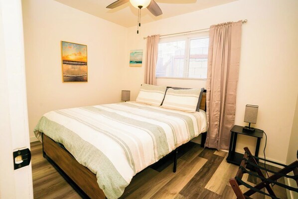 Un dormitorio con cama, ventilador de techo, ventana con cortinas y una mesita de noche pequeña.