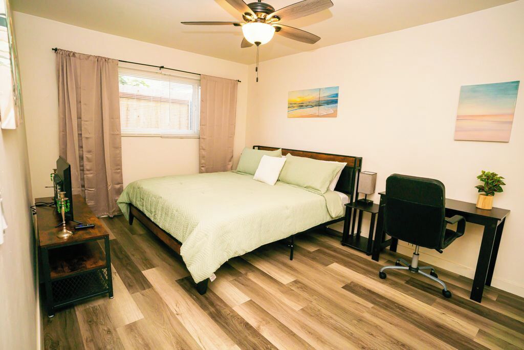 A bedroom with a bed, a desk, a chair, a ceiling fan, and a window with curtains.