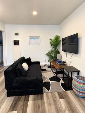 Uma sala de estar moderna com um sofá preto, uma TV de tela plana na parede, uma mesa de centro de madeira, uma planta em vaso e uma cesta colorida de palha.