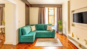 Un canapé d’angle vert avec des coussins blancs, un plancher en bois, un grand téléviseur à écran plat fixé au mur et une plante en pot.