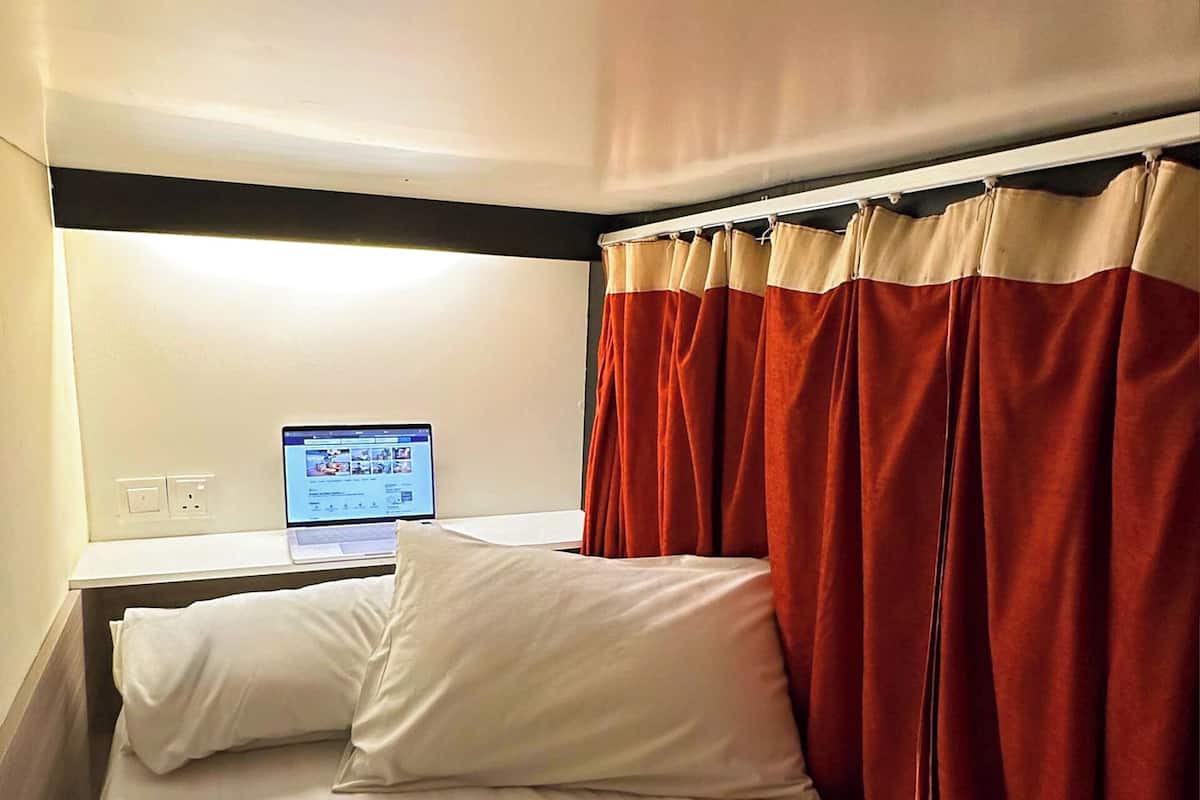 A hotel room with a bed, a laptop on a bedside table, and red curtains.