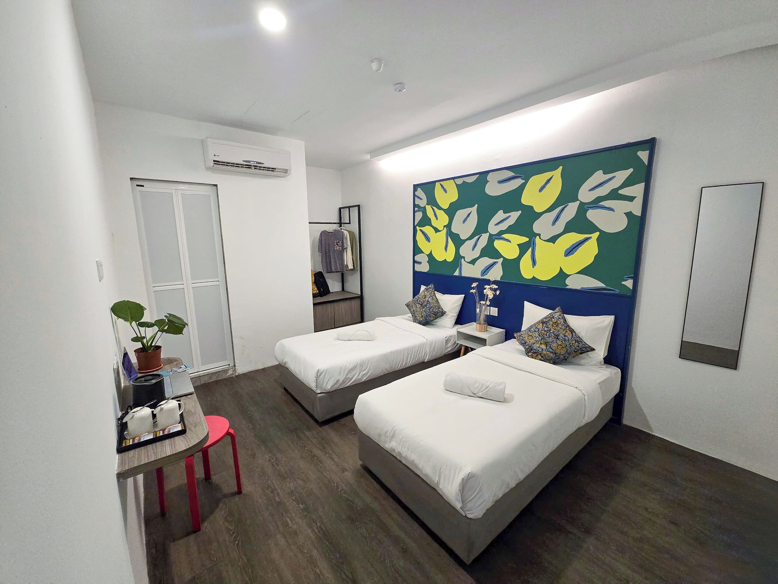 Habitación de hotel con dos camas individuales, un escritorio con silla, una planta en maceta y un amplio adorno mural.