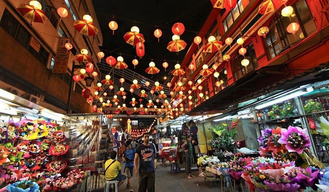 A market with stalls selling flowers and other goods, under a canopy of hanging lanterns.